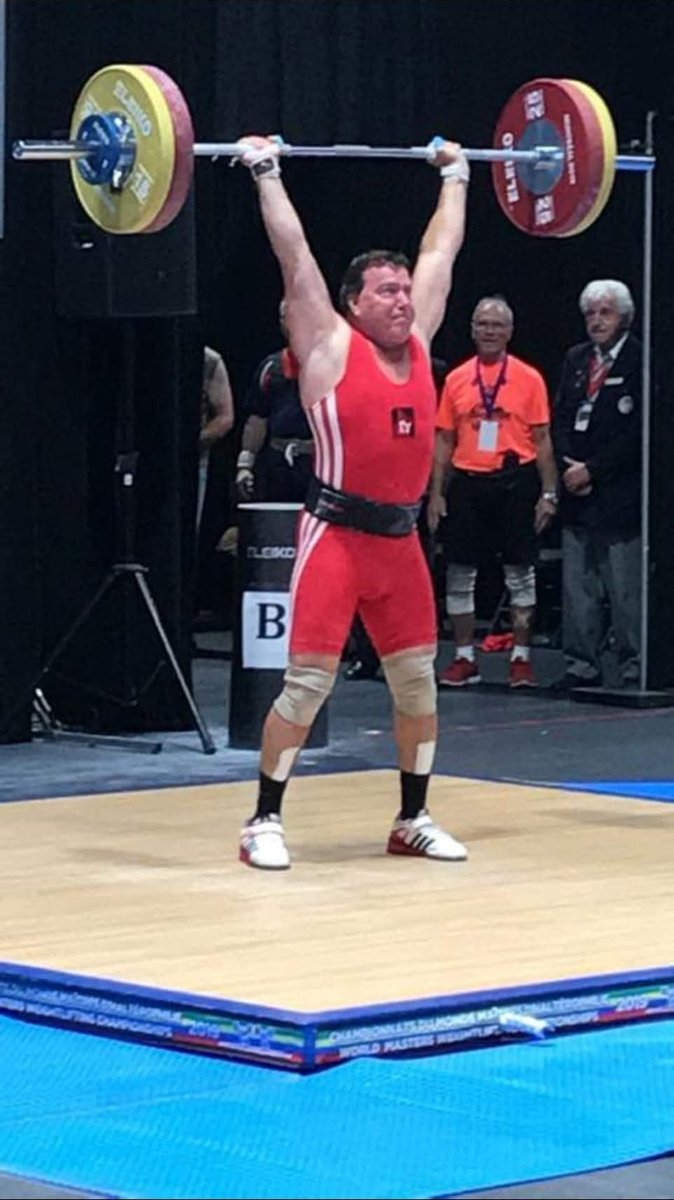 Masters weightlifter André Boutin locking out a jerk overhead in a red singlet on the competition platform at the 2019 World Masters Weightlifting Championships.