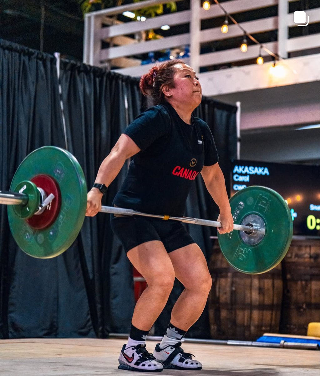 Masters weightlifter Carol Akasaka pulling a snatch in a black Canada team shirt, her name showing on the competition scoreboard behind her.