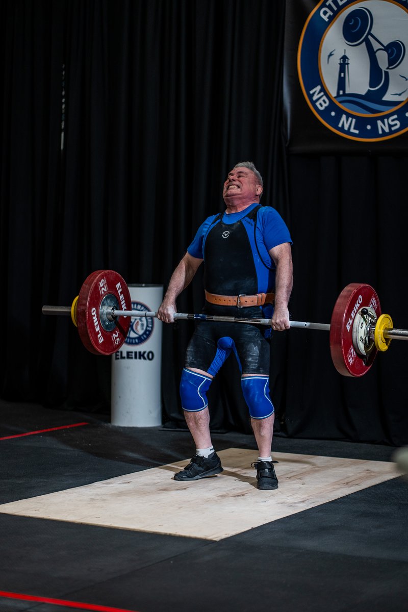 Masters weightlifter Greg Doucett driving through the pull of a clean, teeth gritted with effort, competing in blue at an Atlantic championship meet.