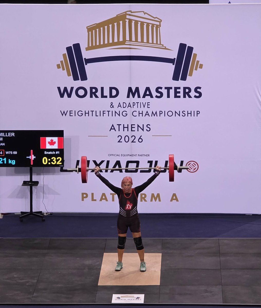 Masters weightlifter Jill Miller holding a snatch locked out overhead on Platform A at the 2026 World Masters and Adaptive Weightlifting Championship in Athens, competing in a black Canada singlet, with the scoreboard beside her showing her name, the Canadian flag and her W75 category.