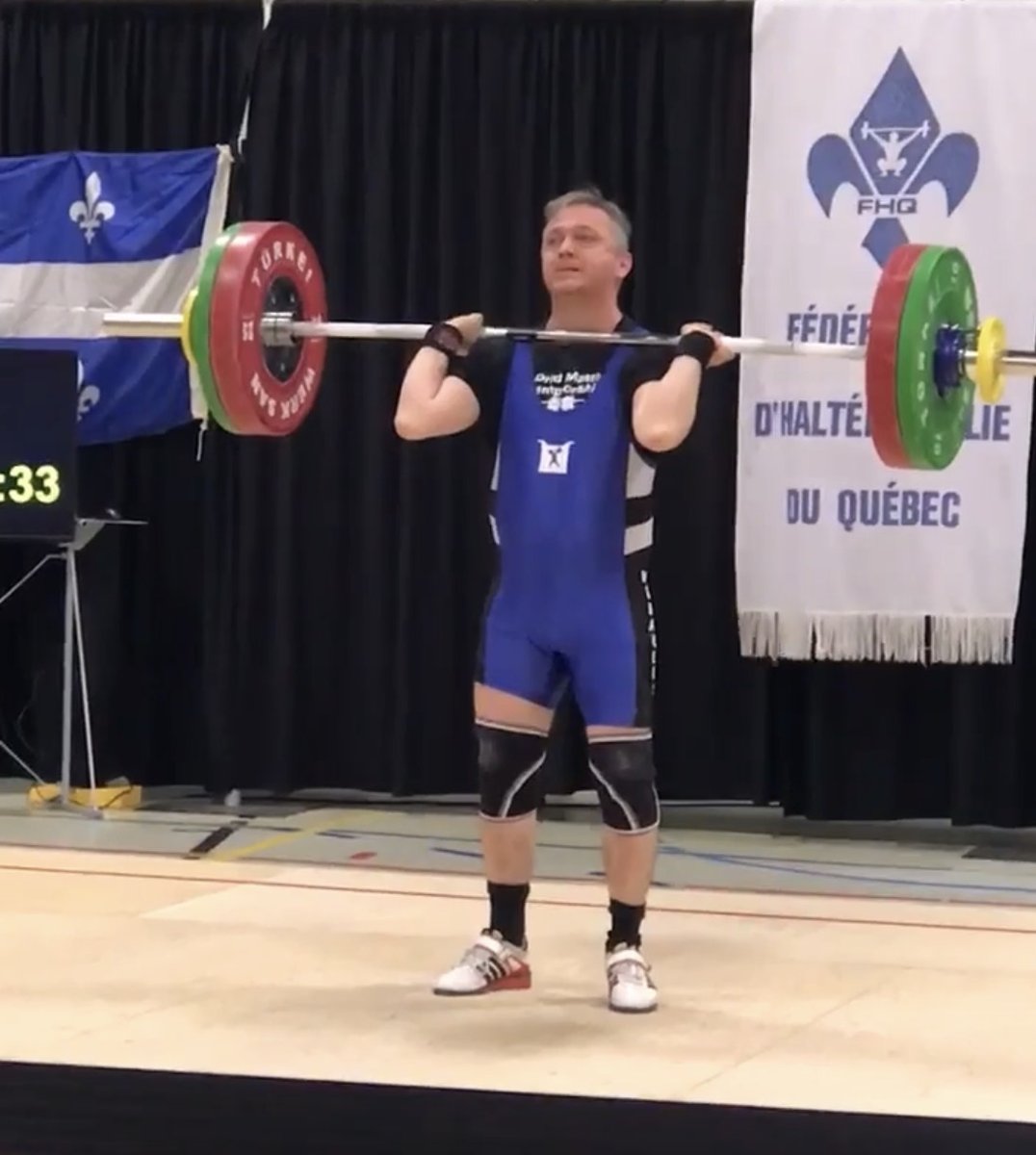 Masters weightlifter Mario Robitaille standing up from a clean with the barbell racked on his shoulders, competing in a blue singlet beneath the Fédération d'haltérophilie du Québec banner.