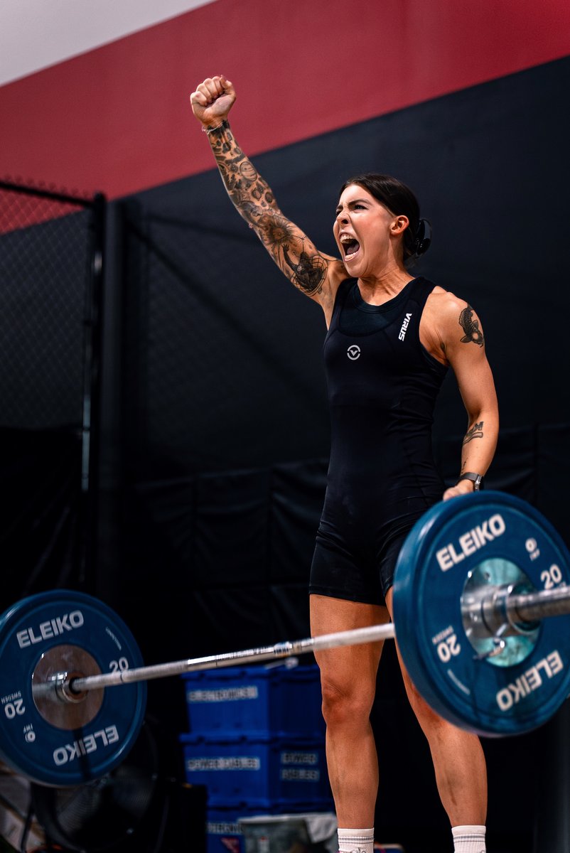 Masters weightlifter Paige Hunter celebrating with a raised fist and a shout after a successful lift, the loaded barbell on the platform in front of her.
