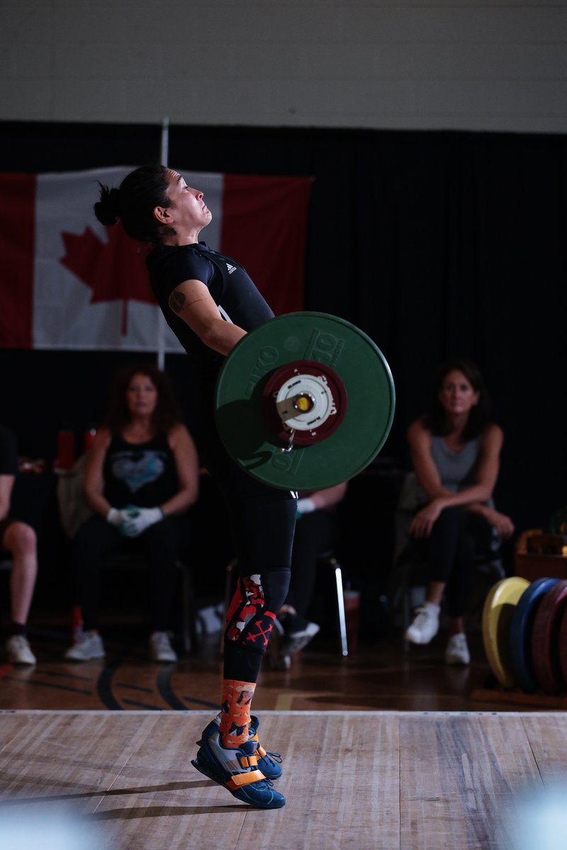 Masters weightlifter Sandra Olivos finishing the pull on a lift in front of a Canadian flag, spectators watching from behind the platform.